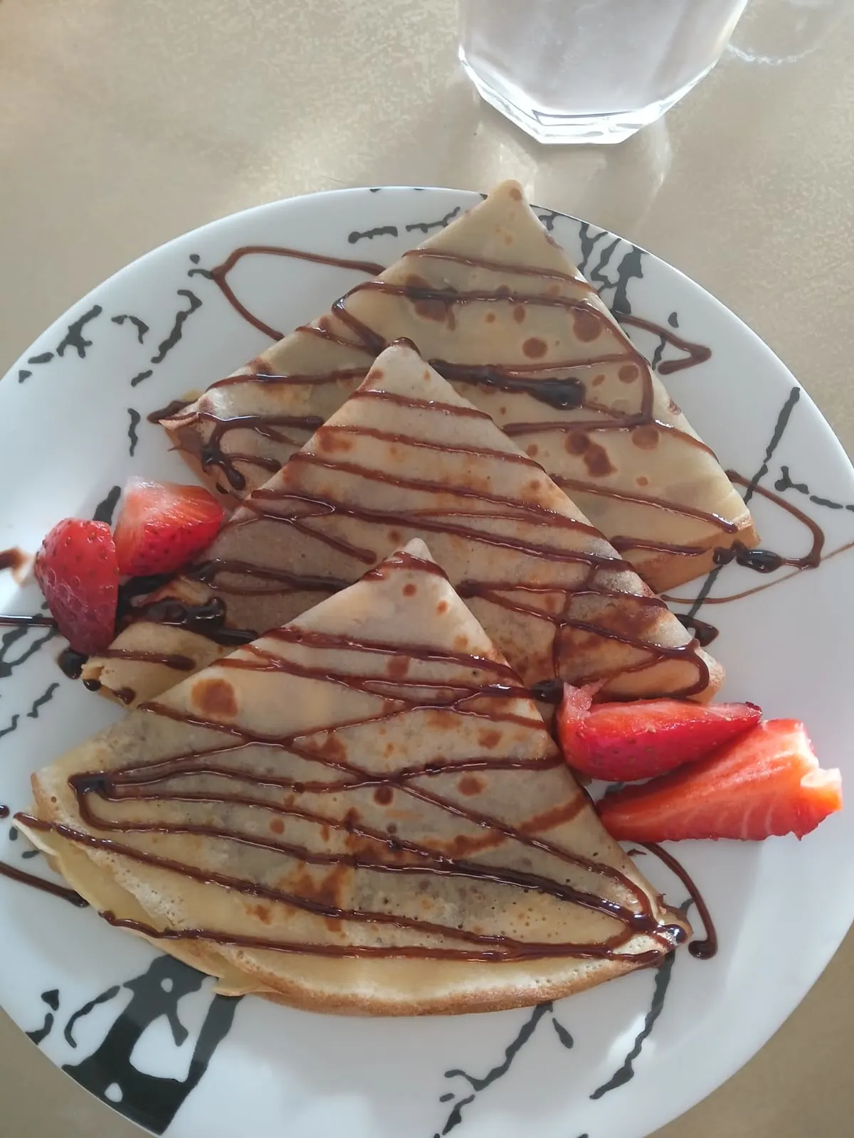 Thin, golden-brown crepes drizzled with chocolate sauce, garnished with fresh strawberries on a decorative plate.